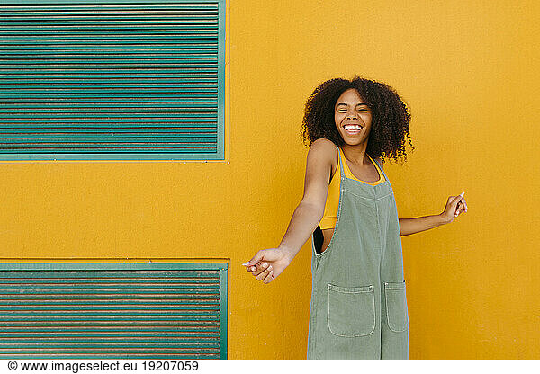 Portrait of happy young woman wearing overalls in front of yellow wall