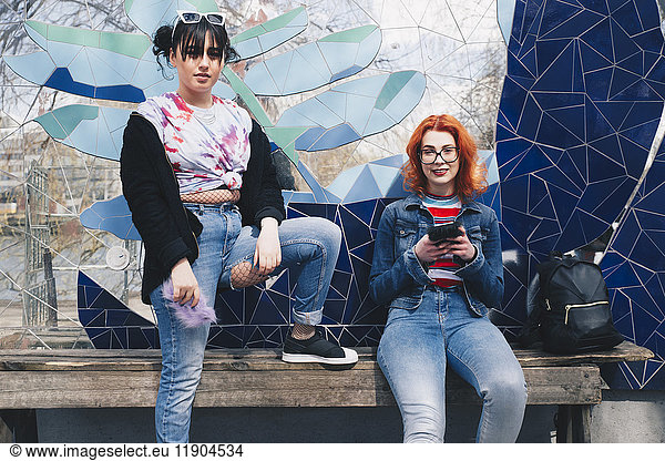 Portrait of hipster female friends holding mobile phones against mosaic wall