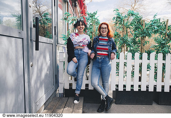 Portrait of hipster female friends standing against fence by building