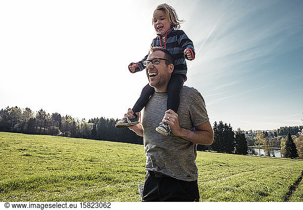 Portrait of laughing father carrying little daughter on his shoulders on a meadow