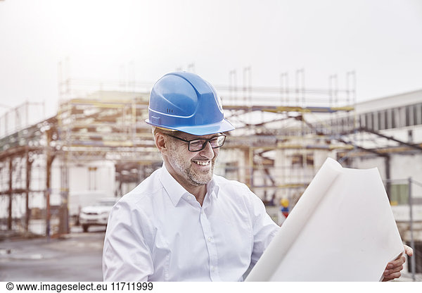 Portrait of laughing man wearing blue hart hat looking at construction plan