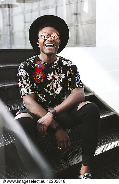 Portrait of laughing man wearing hat  sunglasses and black t-shirt with floral design sitting on stairs