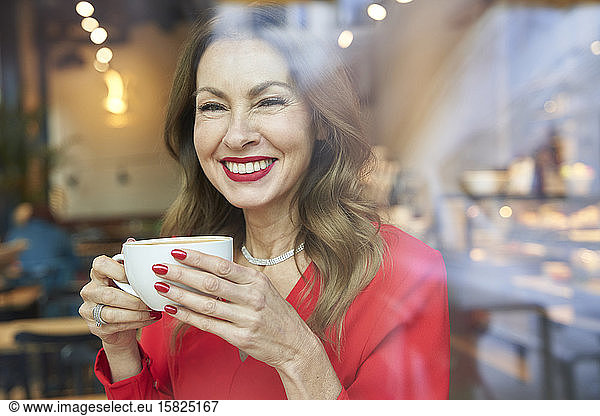 Portrait of laughing mature woman with cup of coffee behind windowpane