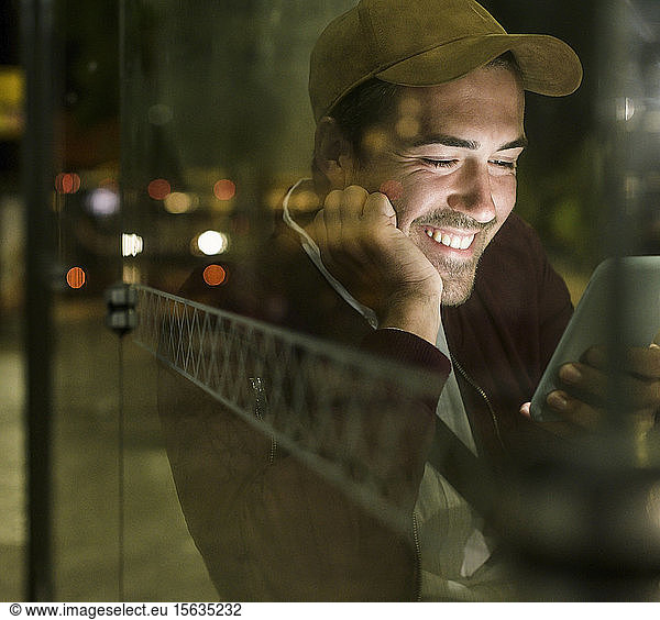Portrait of laughing young man sitting at bus stop by night using cell phone  Lisbon  Portugal