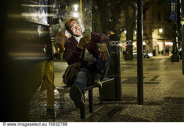 Portrait of laughing young man sitting at bus stop by night using smartphone  Lisbon  Portugal