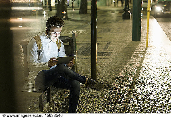 Portrait of laughing young man sitting at bus stop using digital tablet and earphones by night  Lisbon  Portugal