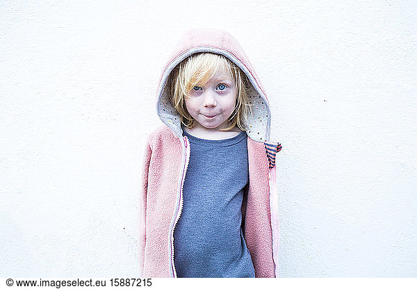 Portrait of little blond girl wearing pink hooded jacket in front of white wall