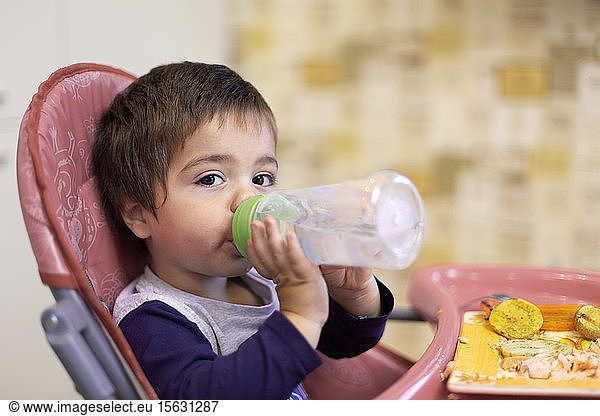 Portrait of little boy sitting on high chair drinking water from flask