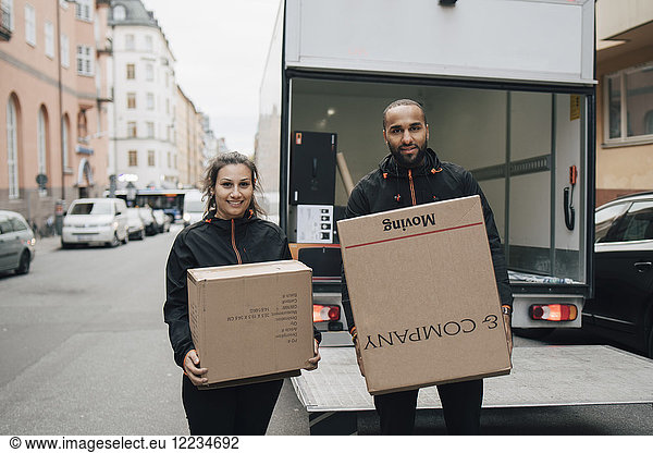 Portrait of male and female workers carrying boxes while standing against delivery van in city