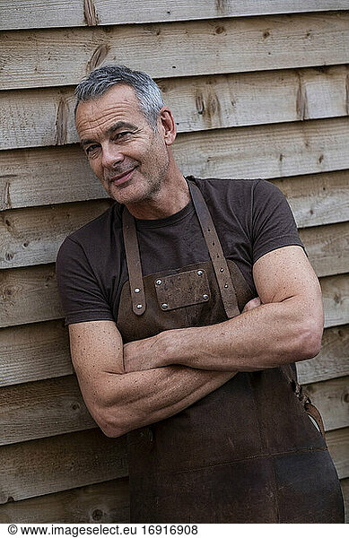 Portrait of male barista with short grey hair  arms folded  leaning against wooden wall.