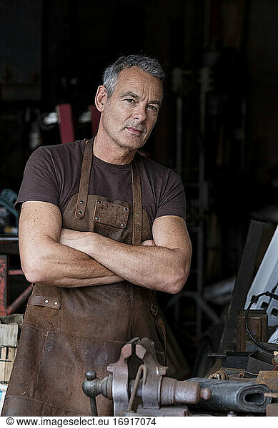 Portrait of male barista with short grey hair  wearing brown apron  arms folded  looking at camera.