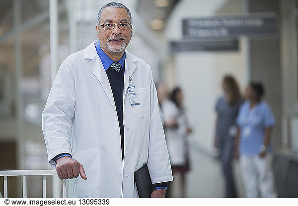 Portrait of male doctor standing in corridor while female coworkers discussing in background