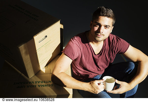 Portrait of man holding coffee cup sitting by cardboard boxes at new home