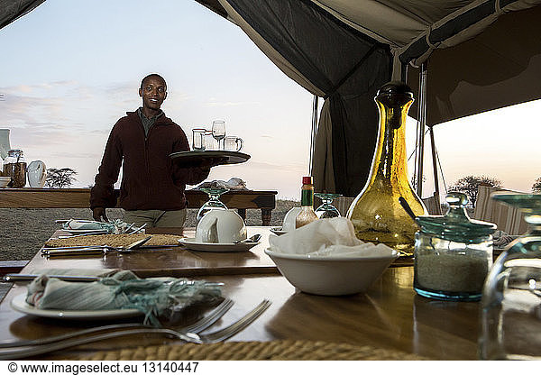 Portrait of man holding tray with drinking glasses by dining table at Serengeti National Park