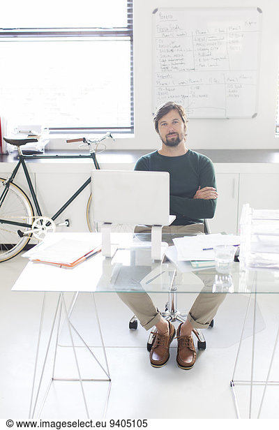 Portrait of man sitting with arms crossed behind glass desk in modern office  bicycle and whiteboard in background