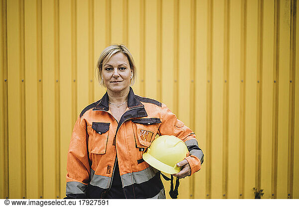Portrait of mature female construction worker with hardhat against yellow metal wall