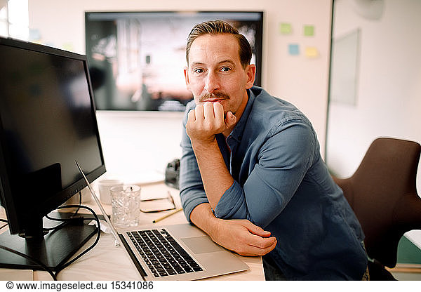 Portrait of mature male professional with hand on chin sitting at office desk