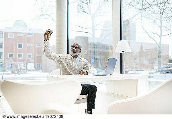 Portrait of mature man with laptop sitting at desk taking selfie with smartphone