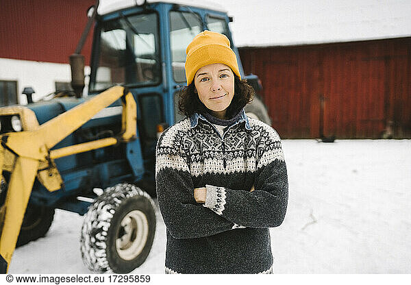 Portrait of mature woman standing with arms crossed against tractor during winter