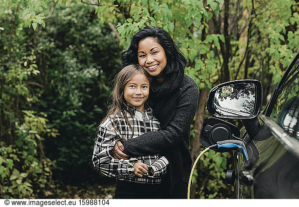 Portrait of mid adult woman with daughter by electric car at charging station