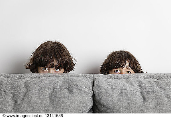 Portrait of playful brothers hiding behind sofa against wall at home
