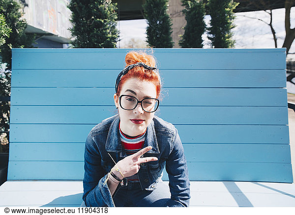 Portrait of redhead young woman gesturing peace sign while sitting on bench