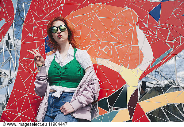 Portrait of redhead young woman wearing sunglasses while gesturing peace sign against mosaic wall