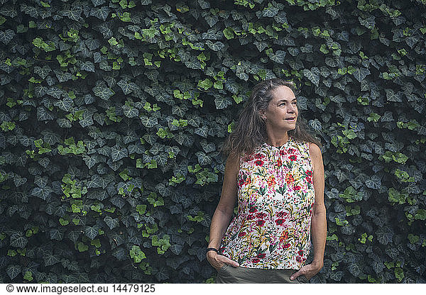Portrait of relaxed mature woman standing in front of wall overgrown with ivy wearing top with floral design