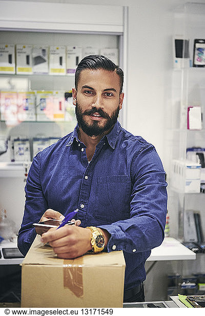 Portrait of salesman writing on box while holding mobile phone in electronics store