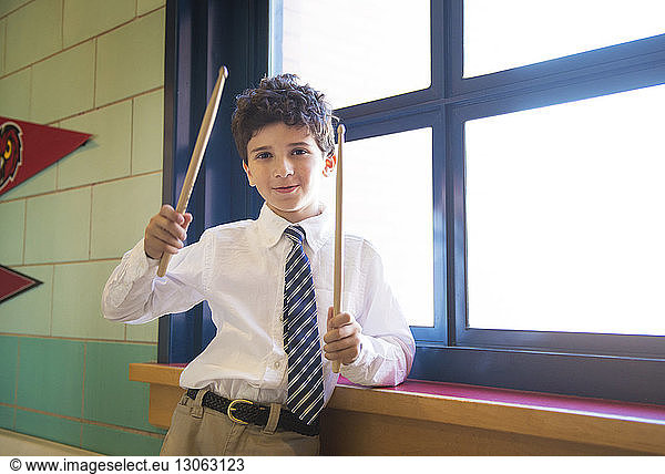 Portrait of schoolboy holding drumsticks while standing at window