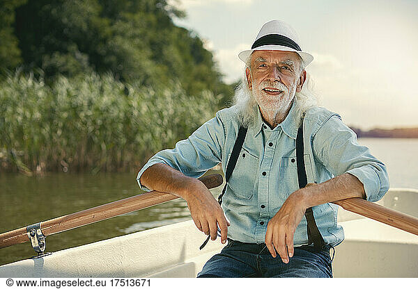 Portrait of senior man sitting in rowing boat on a lake wearing suspenders and summer hat