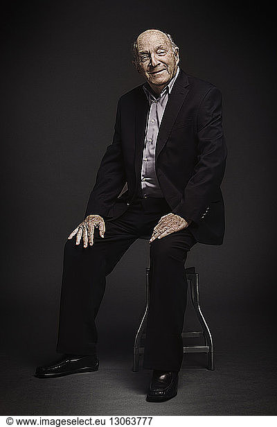 Portrait of senior man sitting on stool against black background