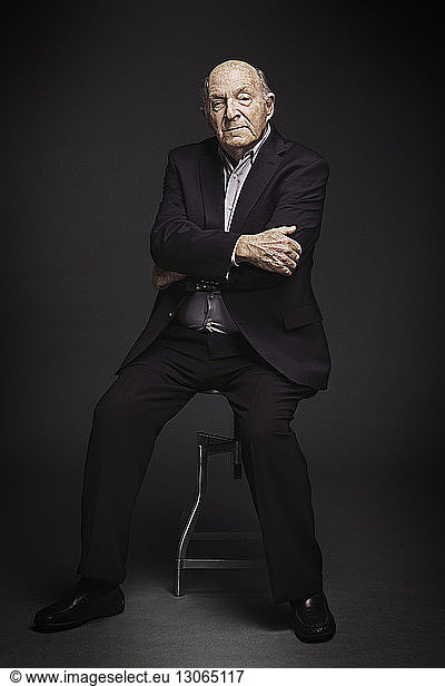 Portrait of senior man with arms crossed sitting on stool against black background