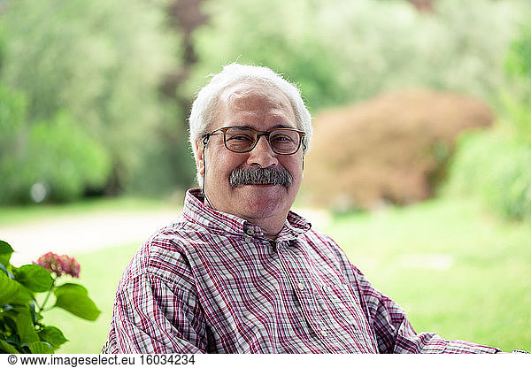 Portrait of senior man with moustache wearing glasses sitting in garden  smiling at camera.