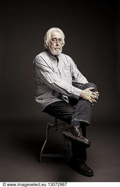 Portrait of senior man with white hair sitting on stool against black background