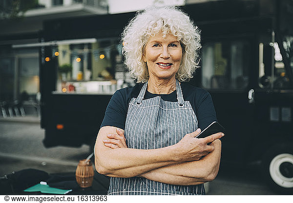 Portrait of senior owner with smart phone standing against food truck in city