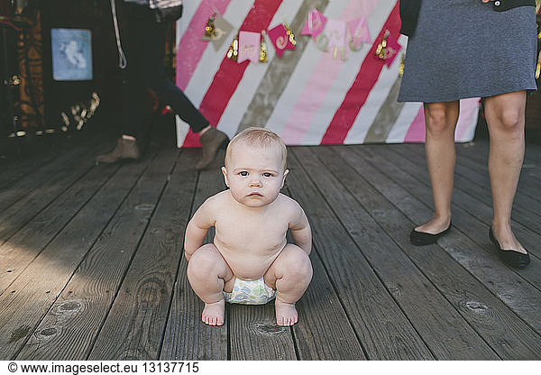 Portrait of shirtless baby girl crouching on floorboard at birthday party with woman standing in background