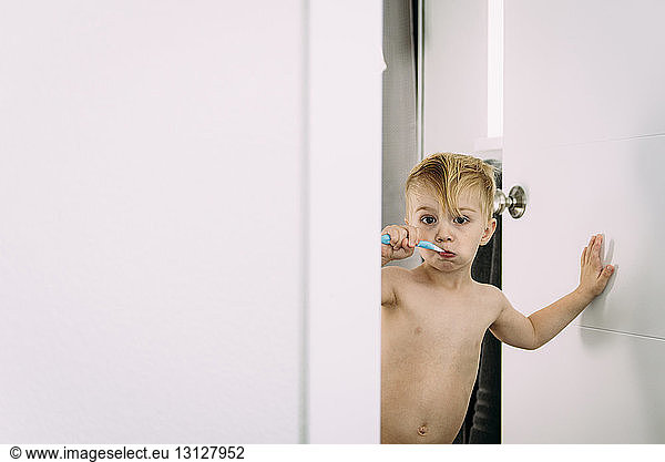 Portrait of shirtless boy brushing teeth while standing at doorway in bathroom