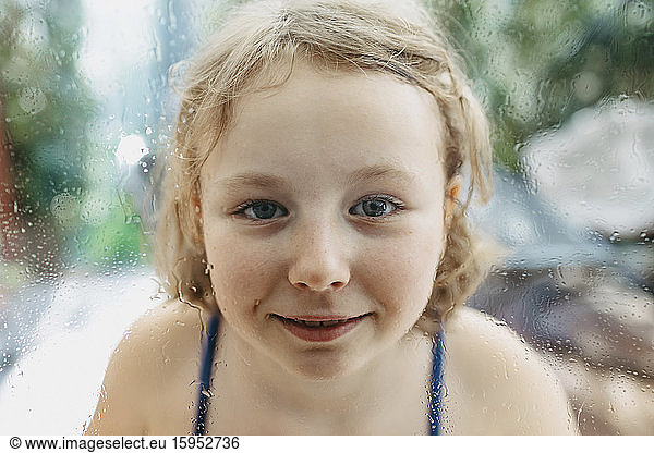 Portrait of smiling blond girl looking through wet windowpane