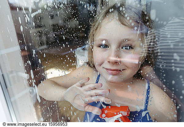 Portrait of smiling blond girl with spray bottle looking through wet window