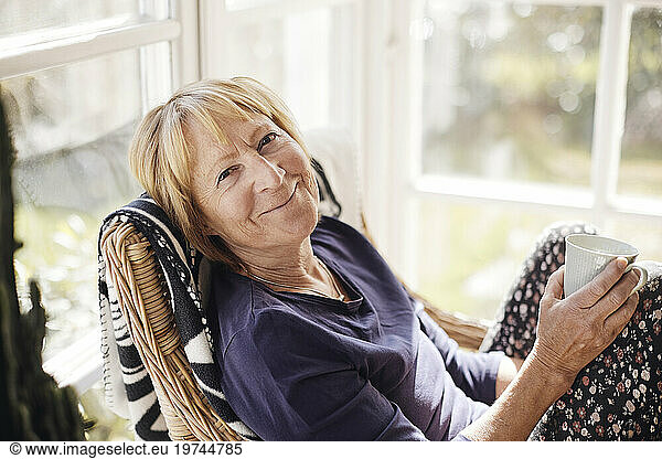 Portrait of smiling blond senior woman with coffee cup sitting on chair at home