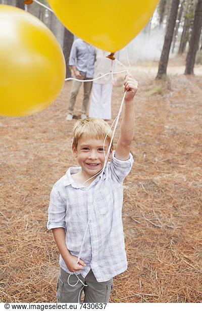 Portrait of smiling boy holding balloons in woods with parents in background