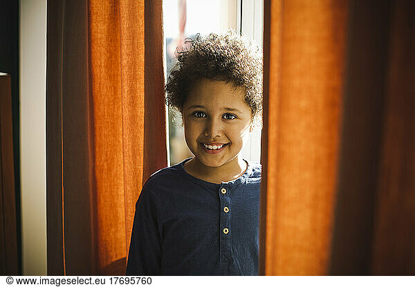 Portrait of smiling boy standing behind curtain at home