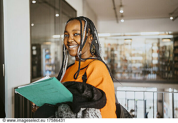 Portrait of smiling braided female student holding jacket and file in university