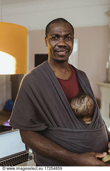 Portrait of smiling businessman carrying toddler son in baby carrier at home