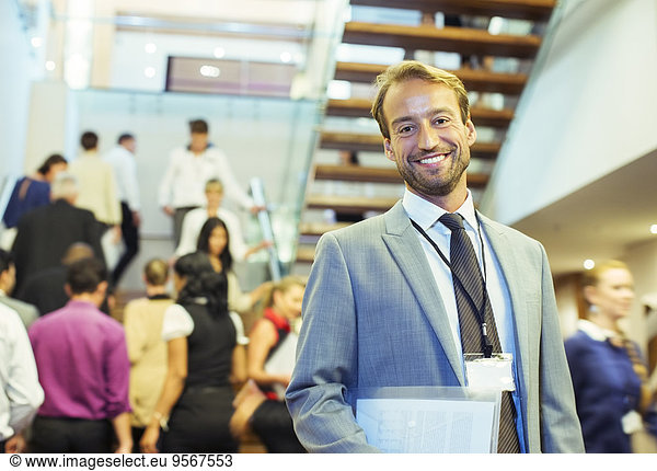 Portrait of smiling businessman holding file  standing in crowded lobby of conference center
