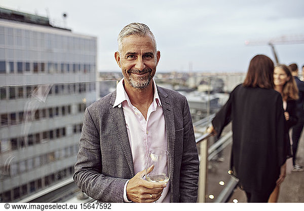 Portrait of smiling businessman holding wineglass with coworkers in background on terrace