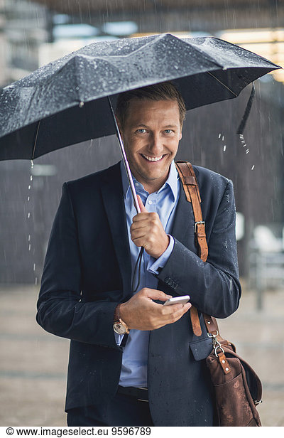 Portrait of smiling businessman using smart phone in city during rainy season
