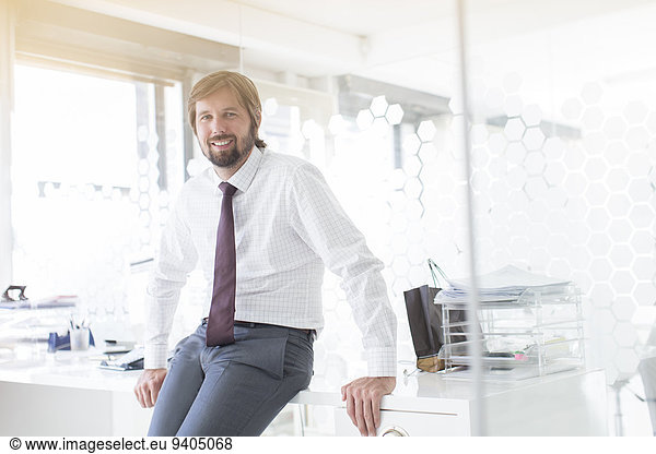 Portrait of smiling businessman wearing shirt and tie leaning on desk in office