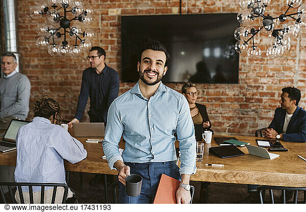 Portrait of smiling businessman with coffee cup and file standing while colleagues discussing in background